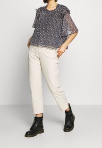 Blouse à imprimé floral avec manches à volants, fond bleu marine, et couleurs incluant le rose et le vert. Associée à un pantalon clair et des bottes noires.