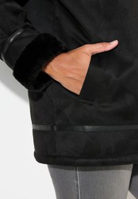 Giacca nera con polsini in pelliccia sintetica e una texture liscia. Presenta una tasca laterale e una striscia di pelle orizzontale vicino all'orlo.