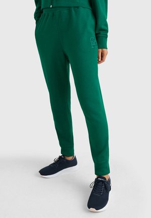 Personne portant un pantalon de jogging vert foncé avec une ceinture élastique et un logo brodé, associé à des baskets bleu marine avec des semelles blanches.