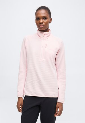 Kvinde iført en lyserød langærmet halv-lynlås-pullover og sorte bukser, stående mod en ensfarvet hvid baggrund.
