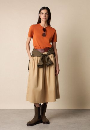 Frau steht, trägt ein orangefarbenes Kurzarmoberteil, einen beigen Midirock mit seitlichen Knöpfen, einen braunen Gürtel, eine gebundene olivgrüne Jacke, braune Stiefel und eine Sonnenbrille am Halsausschnitt.