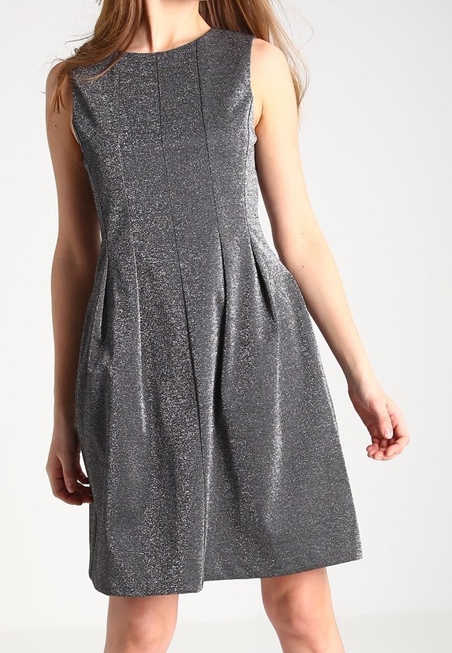 Robe ligne A sans manches argentée pailletée avec plis, longueur genou, portée par une femme debout sur un fond uni.
