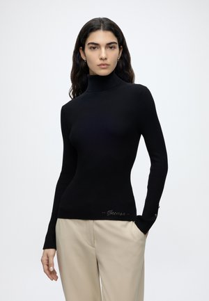 Mujer con cabello largo y oscuro, vistiendo un suéter negro ajustado de cuello alto y pantalones beige, de pie frente a un fondo claro y liso.
