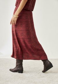 Falda maxi de color rojo con un estampado, con una silueta fluida, combinada con botas marrones a media pantorrilla con tacón cuadrado. Textura de tela suave.