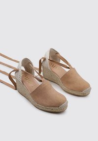 Zapatos de cuña de gamuza color marrón claro con correas de tela trenzada en el tobillo, que cuentan con una suela de yute trenzado y una punta redonda suave. Acentuados con un logo dorado.