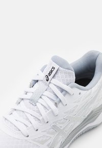 Witte sportieve schoen met een bovenwerk van mesh, lichtgrijze accenten en een klassiek vetersysteem. Bevat een logo op de tong en een gestructureerde zool.