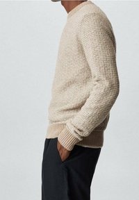 Mann in einem beigen strukturierten Strickpullover mit gerippten Bündchen und Saum, kombiniert mit schwarzen Hosen, steht mit den Händen in den Taschen.