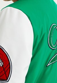 Vihreä ja valkoinen koripalloteemainen takki, jossa on brodeerattuja yksityiskohtia, mukaan lukien punainen koripallopatchi hihassa ja vihreä numero selässä.