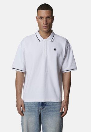 Homme portant un polo blanc à manches courtes avec liseré noir et petit logo sur la poitrine gauche, assorti à un jean bleu clair, debout en pose neutre.