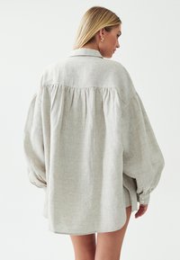 Světle béžová oversized košile vyrobená z lnu, s plisovaným zadním dílem, velkými rukávy, knoflíkovými manžetami a zakulaceným lemem.