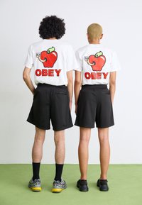 Fehér pólók piros alma grafikával és "OBEY" felirattal, fekete nadrágokkal párosítva. A modellek hátukkal egymás felé állnak, bemutatva a dizájnokat.