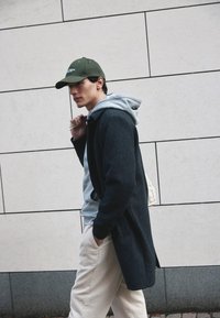 Dunkelgrauer Mantel über einem hellgrauen Hoodie, kombiniert mit beigen Cordhosen. Eine grüne Mütze rundet das Outfit ab. Einfache Hintergrund.