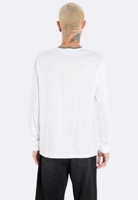 Camicia bianca a maniche lunghe con collo rotondo, con bordi neri sul collo. Indossata con pantaloni in denim scuro, che mostrano una vestibilità comoda.