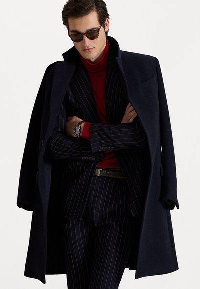 Námornícky kabát nosený cez červený rolák a prúžkovaný oblek. Má úzky strih, kontrastné textúry a okrúhle slnečné okuliare s tmavými sklami.