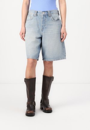 Person iført lyseblå denimshorts og høje mørkebrune læderstøvler, der står mod en hvid baggrund.