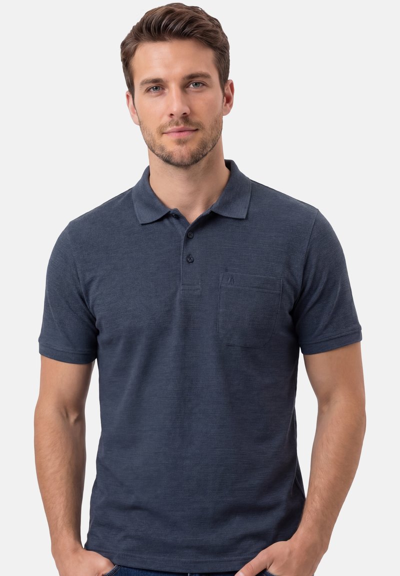 Homme aux cheveux courts bruns portant un polo bleu marine à manches courtes avec une poche avant, debout devant un fond uni.