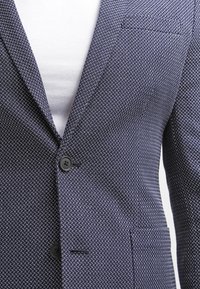 Marinblazer i strukturerad tyg med ett litet vitt geometriskt mönster, enkelknappstängning och bröstficka.