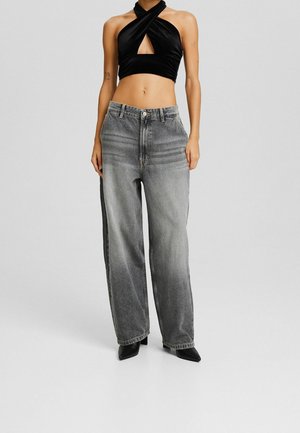 Žena v černém halter crop topu s překříženými pásky a vysokým pasem širokých šedých džínách s černými špičatými botami, stojící před jednobarevným pozadím.