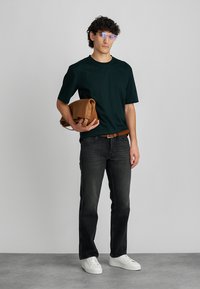 Sötétzöld póló, fekete farmernadrág, barna öv és fehér sportcipő. Egy barna bőrtáskát tart a kezében. A szemüveg modern megjelenést kölcsönöz.