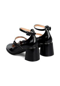 Scarpe Mary Jane in vernice nera, con punta rotonda, cinturini alla caviglia con fibbie e tacchi a blocco spessi su uno sfondo bianco.