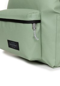 Mochila de nylon verde con un bolsillo frontal con cremallera, acentos negros y una etiqueta. Cuenta con costuras duraderas y una superficie texturizada.