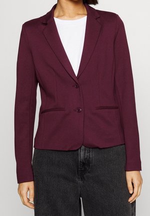 Person iført en tætsiddende mørkelilla blazer med to knapper over en hvid skjorte og mørkegrå jeans mod en ensfarvet baggrund.