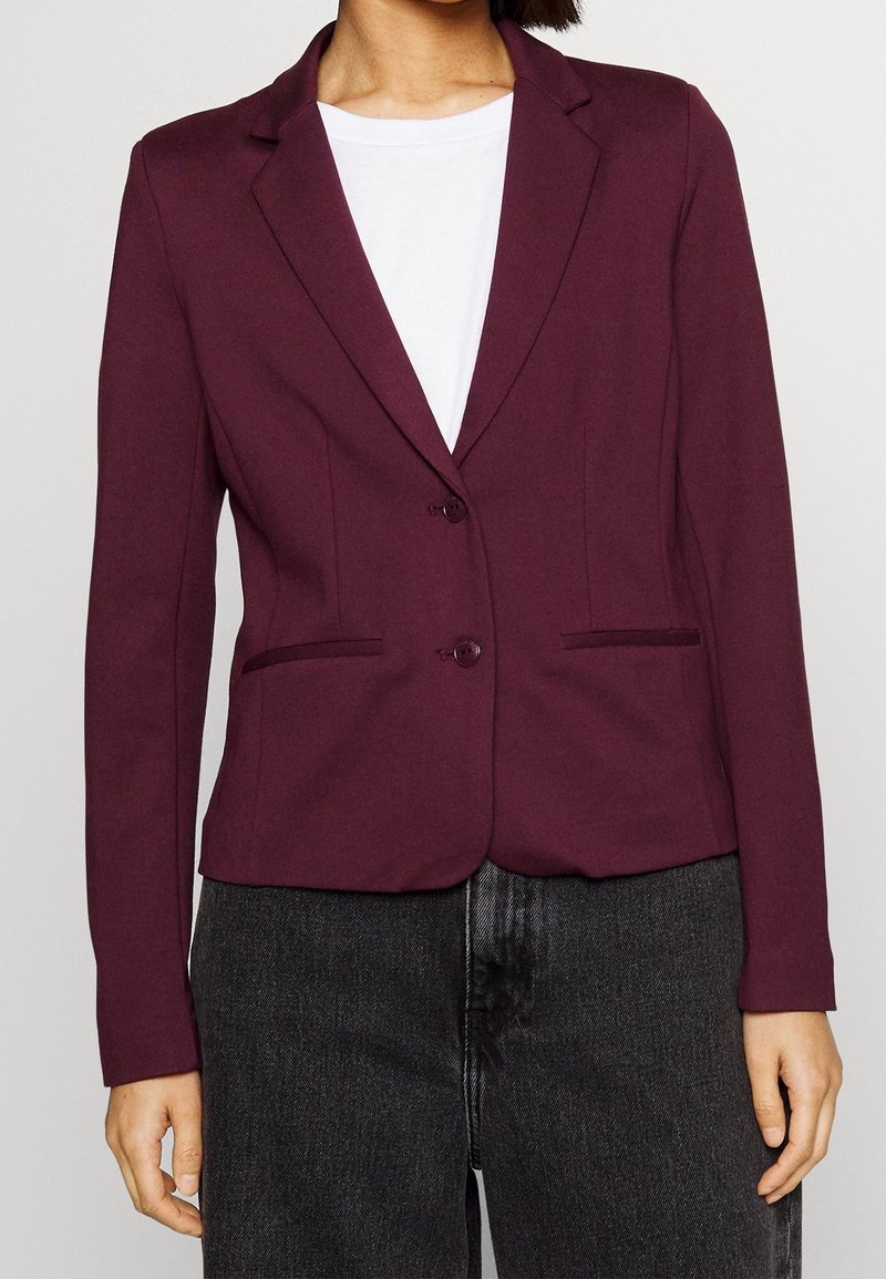 Personne portant un blazer violet foncé ajusté avec deux boutons sur une chemise blanche et un jean gris foncé sur un fond uni.