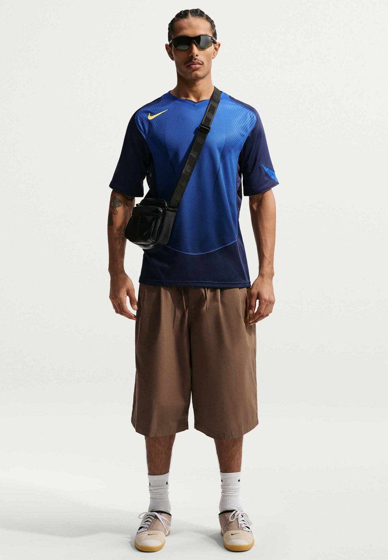 Hombre con camiseta deportiva azul de Nike, pantalones cortos marrones hasta la rodilla, calcetines blancos, zapatillas beige, gafas de sol negras y un pequeño bolso negro bandolera.