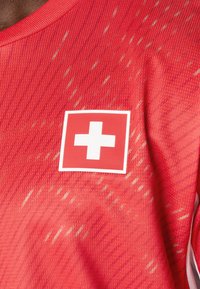 Piros sportpóló texturált mintával és egy fehér svájci zászló folttal a bal mellrészen. Az anyag légáteresztőnek és könnyűnek tűnik.