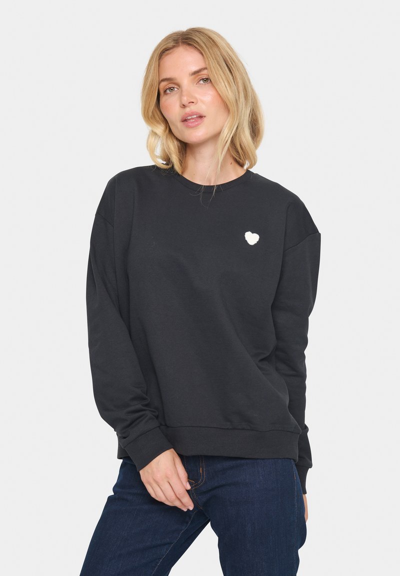 Sudadera negra de manga larga, cuello redondo y logo de corazón blanco texturizado en el pecho. Tejido suave con puños acanalados.