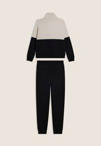 Completo a due pezzi con una maglietta a maniche lunghe color beige e nero a blocchi di colore e pantaloni jogger neri su uno sfondo neutro.