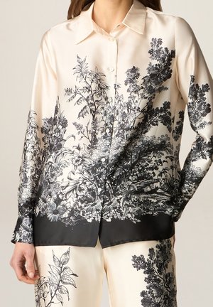 Femme portant une chemise et un pantalon en soie couleur crème ornés d'un imprimé botanique noir détaillé, comprenant des fleurs, des feuilles et des arbres.