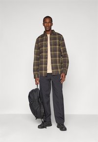 Man in een groen en bruin geruite overhemd, beige T-shirt, zwarte cargobroek, zwarte sneakers, met een zwarte rugzak aan het handvat.