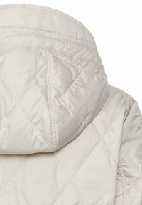 Veste matelassée couleur crème avec capuche rembourrée présentant des coutures en forme de losange et des détails froncés à l'épaule.