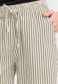 Pantalons légers en tissu beige avec des rayures verticales noires. Dotés d'une taille élastique avec cordon de serrage et de poches latérales.