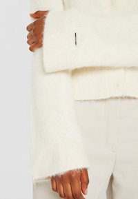 Cardigan blanc et duveteux en tricot avec un col côtelé et une fermeture à boutons. Il présente des manches larges avec des ourlets bruts et un petit accent métallique.