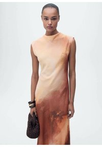 Mouwloze jurk in gradient tinten van perzik en roest, met een abstract aquarelpatroon. Draagt een geweven donkere handtas en armbanden.