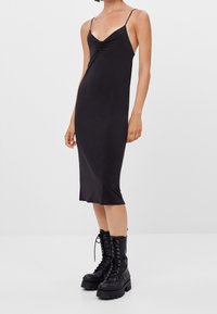Robe noire midi avec une silhouette ajustée, des bretelles spaghetti et un détail froncé au niveau de la poitrine. Assortie avec des bottes de combat noires.