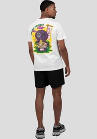 Bílé bavlněné tričko s barevným karikaturou na zádech, znázorňující fialovou postavu s ramenem a dekorativními prvky. Černé šortky.