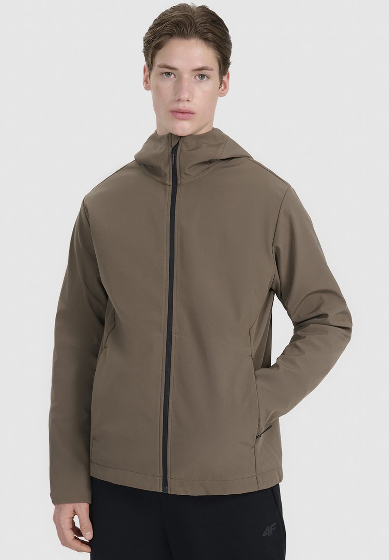 Jeune personne portant une veste à capuche marron avec fermeture éclair sur le devant et un pantalon noir, debout avec une main dans une poche devant un fond clair.