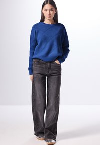 Blå stickad tröja med en texturerad diamantmönster, tillsammans med högmidjade svarta vida jeans och metalliska silverskor.