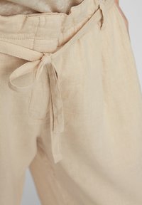 Beżowe lniane spodnie z widocznym paskiem w talii, regulowanym wiązaniem i kieszenią boczną. Materiał wydaje się lekki i lekko teksturowany.