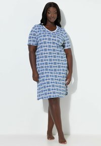 Mujer de pie descalza, con un vestido a la rodilla azul y blanco con estampado y mangas cortas, sobre un fondo blanco liso.