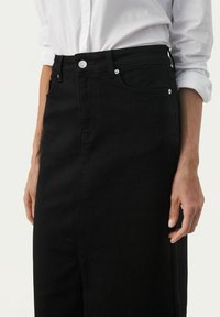 Nainen, jolla on yllään mustat korkeavyötäröiset farkkuhameet ja valkoinen pitkähihainen paita, jonka hihat on käännetty ylös, kädet rentoina sivuilla.