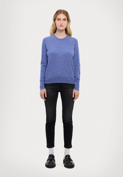 Blå crew-neck tröja i lättviktsmaterial, matchad med mörka jeans och svarta platåskor, stående mot en enfärgad bakgrund.