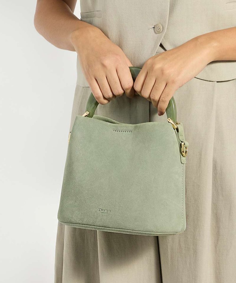 Mains tenant un petit sac à main vert en daim avec une poignée arrondie et des ferrures dorées, porté avec une robe beige boutonnée.