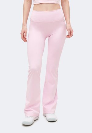 Nő világos rózsaszín, magasderekú, bővülő leggingsben és fehér tornacipőben, sima világos háttér előtt áll.