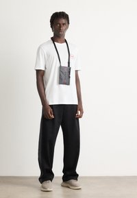 Mann med hvit Hugo T-skjorte, svarte joggebukser, beige sneakers, og en liten grå veske med "HUGO" på en svart reim rundt halsen.