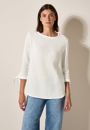 Blouse en lin blanc avec des manches trois-quarts, un col rond et des accents à nouer au niveau des poignets. Tissu texturé avec une coupe décontractée.