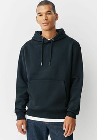 Schwarzer Baumwollhoodie mit einer Vordertasche, verstellbaren Kordeln und Rippbündchen. Getragen über einem weißen T-Shirt und kombiniert mit blauen Jeans.